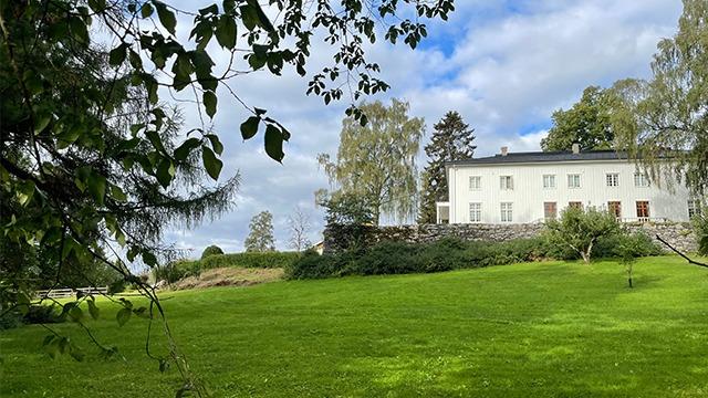 En stor, hvit bygning med vinduer, omringet av grønt gress og trær under en blå himmel.