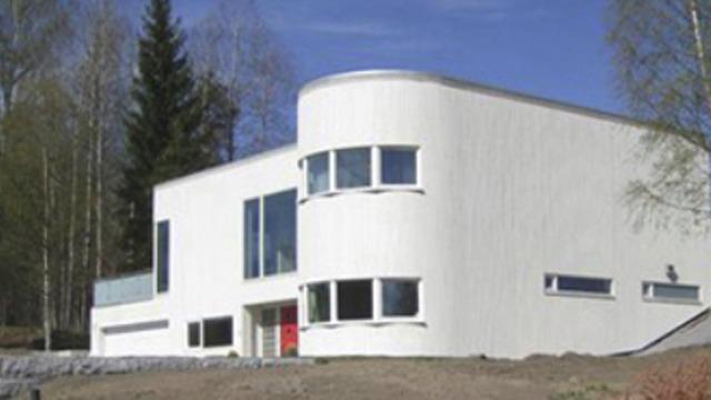 Moderne, hvitt hus med buet fasade og store vinduer, omringet av trær og blå himmel.