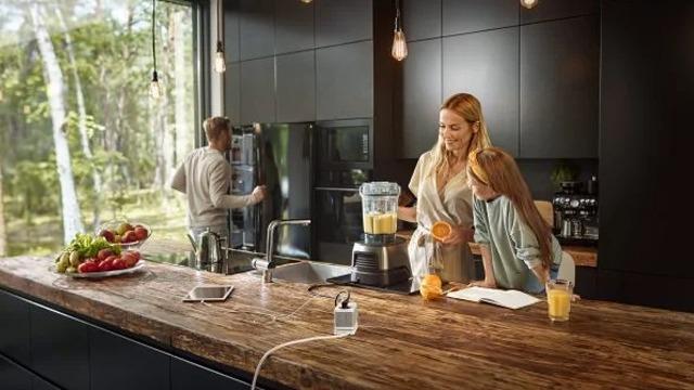 En kvinne lager smoothie i et moderne kjøkken mens en jente ser på, og en mann står ved kjøleskapet.