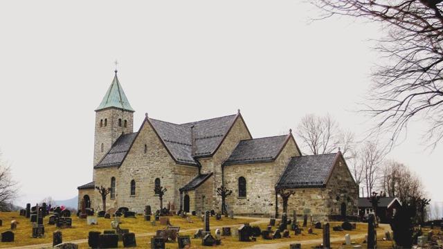 En steinkirke med grønt spir, omgitt av gravsteiner og trær på en bakke.