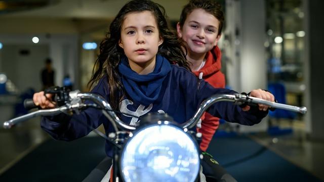 To barn som sitter på en motorsykkel, en jente med langt hår og en gutt med kort hår, begge smilende.
