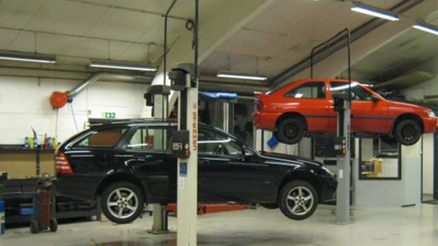 To biler hevet på verksted, en svart stasjonsvogn og en rød hatchback, med verktøy og utstyr i bakgrunnen.