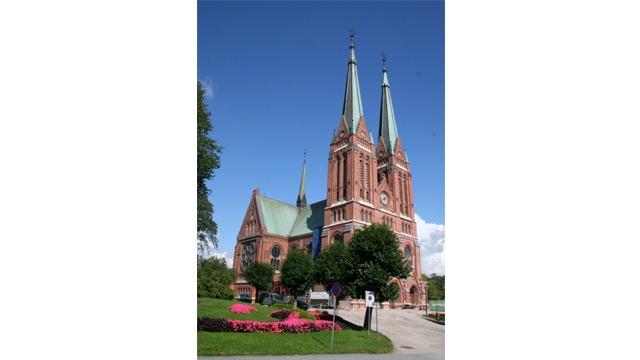 En stor, rød steinkirke med grønne spir og klokketårn, omgitt av trær og blomsterbed.