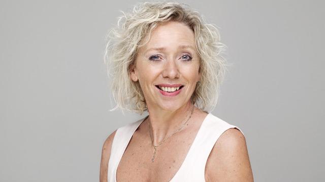 Kvinne med krøllete, blondt hår og smilende ansikt, iført en hvit topp mot en lys grå bakgrunn.