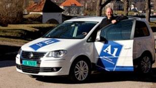 En mann står ved siden av en hvit bil med A1-logo, parkert i et solfylt område.