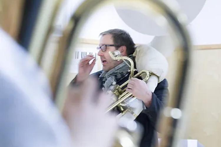 Mann med briller som spiller tuba, iført skjerf, i en øvingslokale med instrumenter i bakgrunnen.