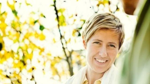 Kvinne med kort hår smiler mot kameraet, omringet av høstfargede trær.