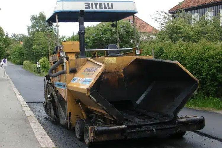 Asfaltleggermaskin i arbeid på en vei, med grøntområde og bygninger i bakgrunnen.