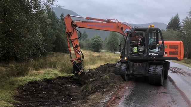 Gravemaskin som graver langs veien i et skogsområde med fjell i bakgrunnen.