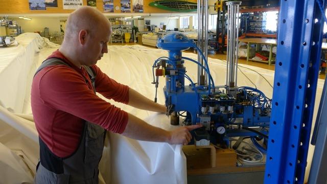 En mann arbeider med en blå maskin i en verksted, omringet av hvite materialer og verktøy.