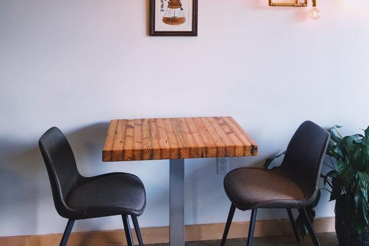Et moderne bord i tre med to stoler, plassert mot en lys vegg med kunstverk og en plante i bakgrunnen.