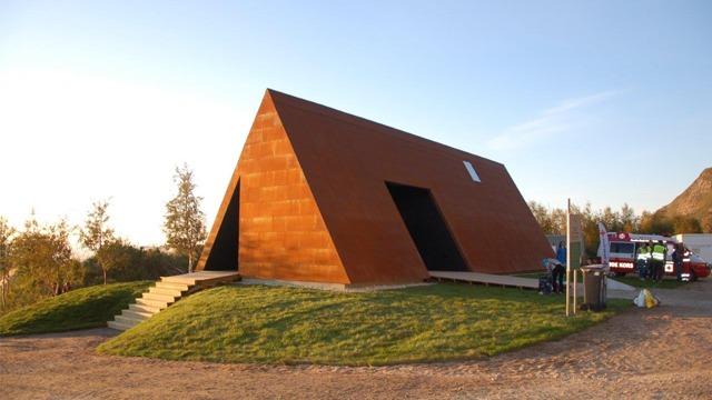 Moderne, trekantet hus med rustfarget fasade, plassert på en grønn bakke under en klar himmel.