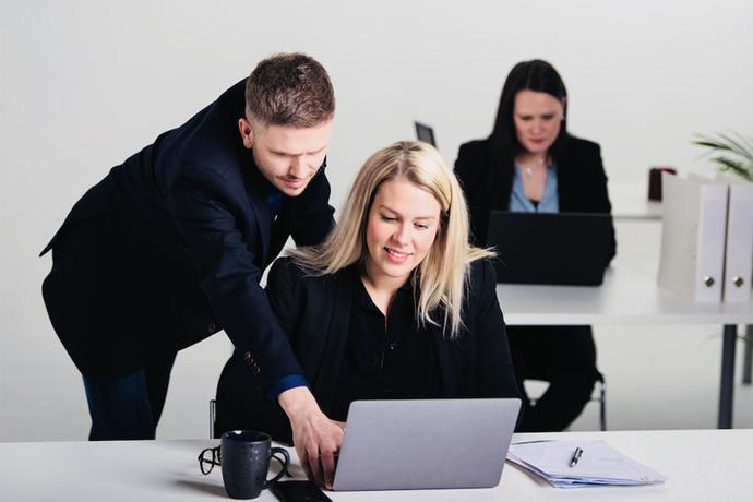 To personer i et kontor, en mann hjelper en kvinne med en laptop, mens en annen kvinne jobber i bakgrunnen.
