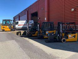 Flere gule gaffeltrucker parkert foran en rød lagerbygning med en lastebil i bakgrunnen.