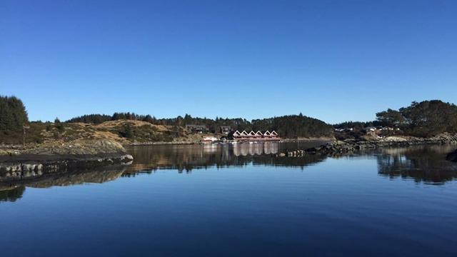 Roelig sjølandskap med speilbilde av bygninger og fjell i bakgrunnen under klar blå himmel.
