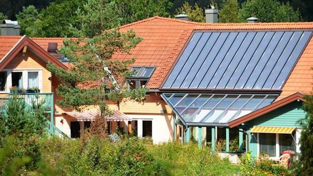 Moderne hus med solcellepaneler på taket og en stor vinterhage omgitt av grønt landskap.