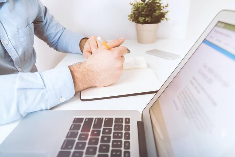 En person med skjorte sitter ved et skrivebord med en laptop, notatbok og en potteplante i bakgrunnen.