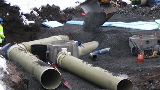 Byggeplass med rør og grus, arbeidere i sikkerhetsklær, snø og utstyr synlig i bakgrunnen.