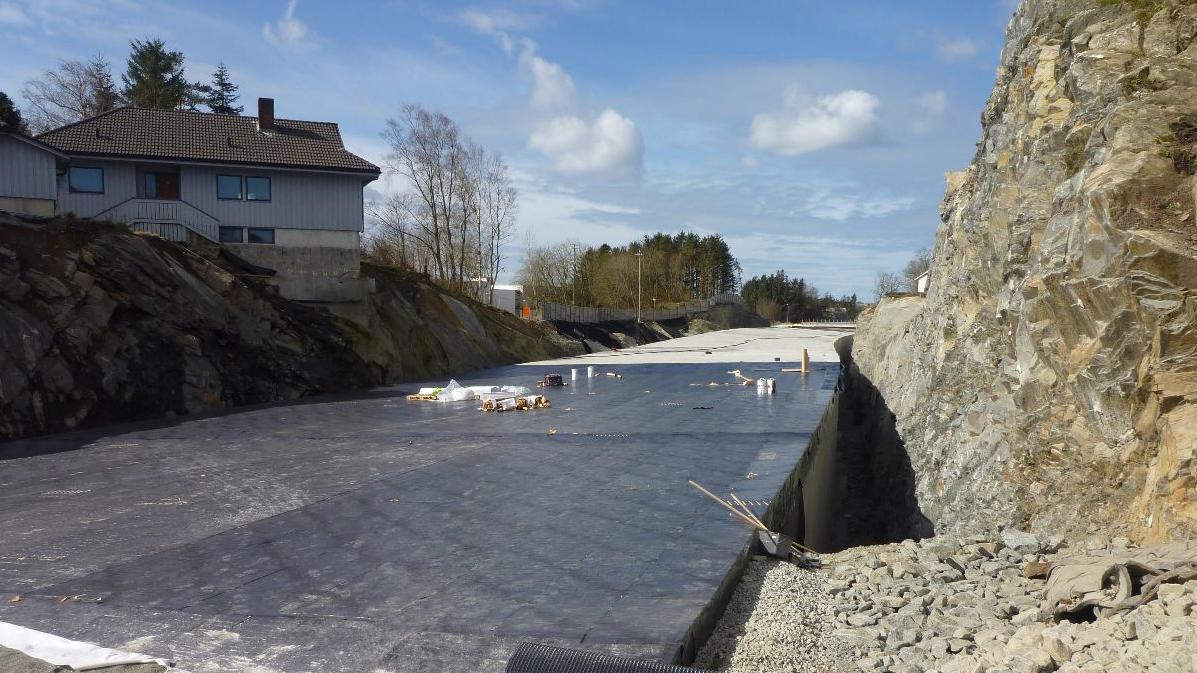 Byggeplass med grus og duk, omgitt av fjell og hus i bakgrunnen under en blå himmel.