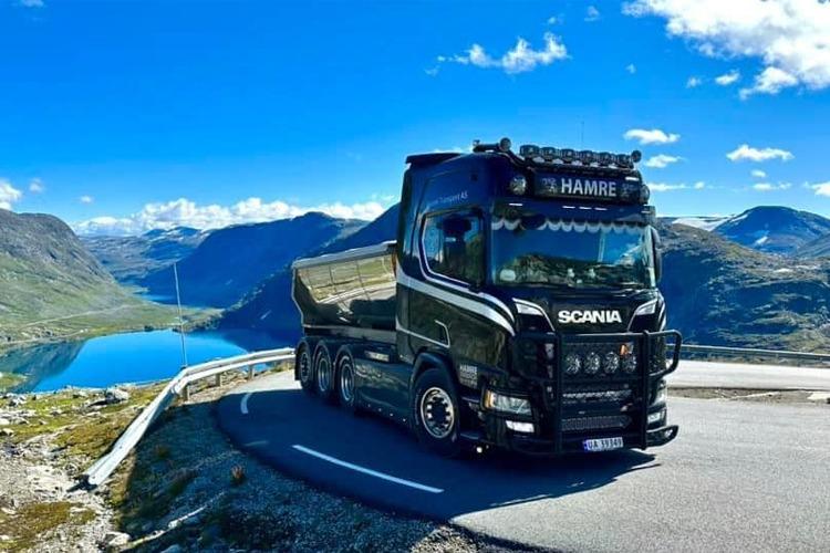 En svart Scania lastebil med tippvogn på en fjellvei med innsjø og fjell i bakgrunnen.