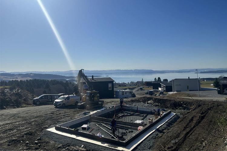 Byggeplass med fundament for hus, gravemaskin og biler, med utsikt over innsjøen og fjellene i bakgrunnen.
