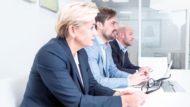Tre personer i et møterom, to menn og en kvinne, som ser på en datamaskin og noterer under et møte.