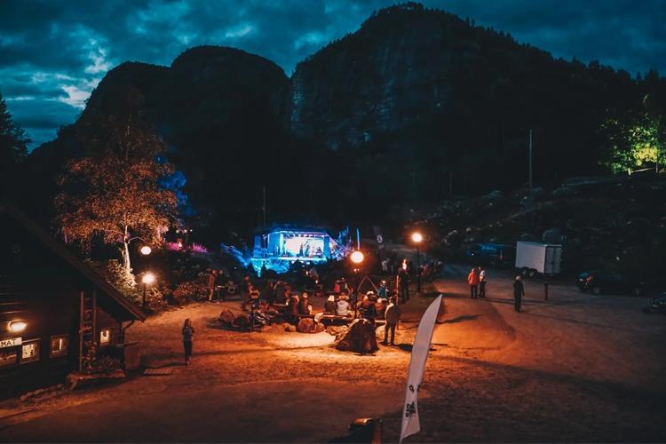 En utendørs konsert med folk som samles rundt scenen i en naturskjønn setting med fjell i bakgrunnen.