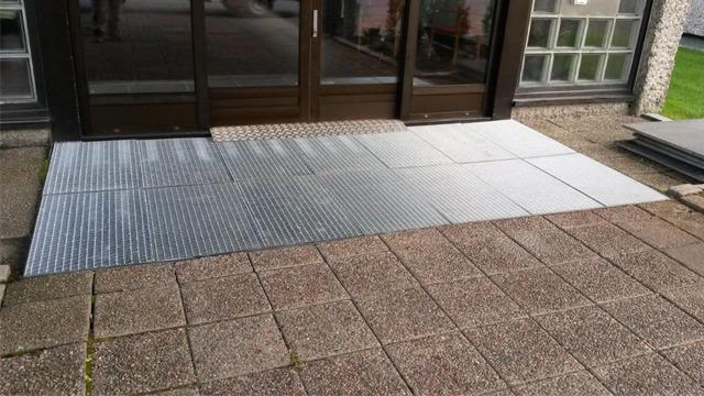 En rampe laget av metallplater foran en inngangsdør, med steinlagt fortau på siden.