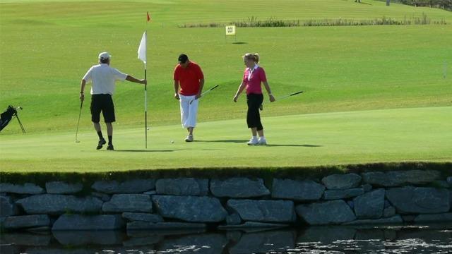 Tre personer spiller golf på en grønn golfbane, med en flaggpinne og vann i bakgrunnen.