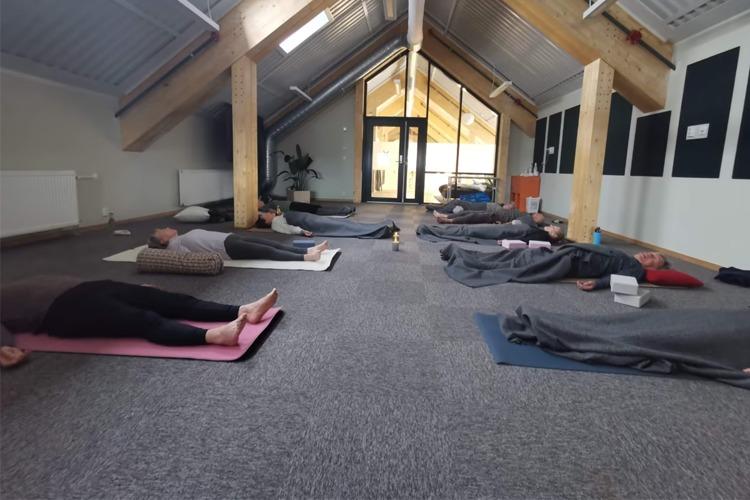 En gruppe mennesker ligger på yogamatter i et rom med skråtak, omgitt av naturlig lys og en avslappende atmosfære.