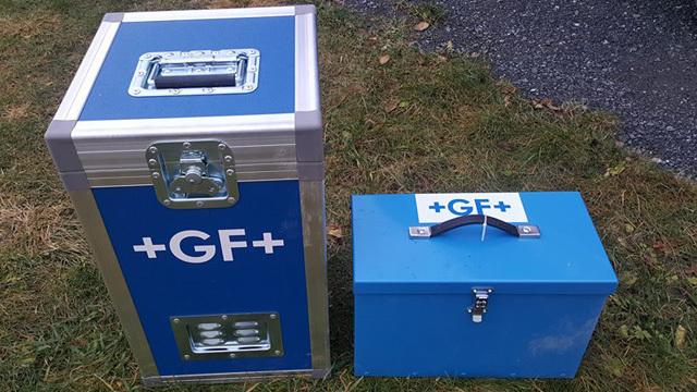 To kasser i blått metall med +GF+ logo, en stor kasse og en mindre kasse, plassert på gress.
