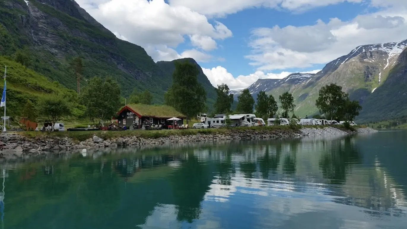 Campingplass ved en innsjø omkranset av fjell og trær, med campingvogner og en hytte i bakgrunnen.