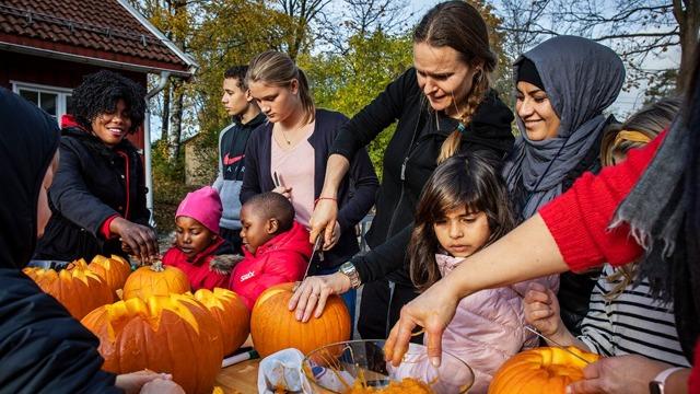 Familier og barn skaper gresskar i en utendørs aktivitet, med smil og samarbeid i en høstlig setting.