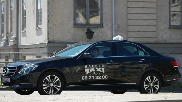 En svart Mercedes taxi med teksten "Halden Taxi" og telefonnummeret 69 21 32 00 står parkert på gaten.