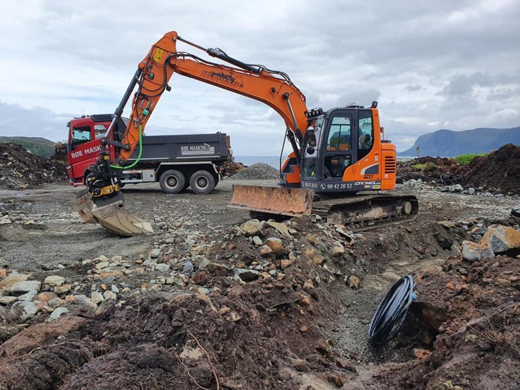 Gravemaskin i arbeid på en byggeplass med lastebil i bakgrunnen og stein og jord rundt.