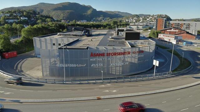 Moderne bygning med glassfasade og stor logo for Åsane Storsenter, omgitt av veier og fjell i bakgrunnen.
