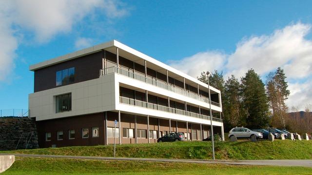 Moderne bygning med flere etasjer, balkonger og parkerte biler på en grønn plen under en blå himmel.