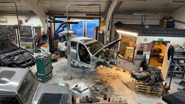 Verksted med flere biler, en bil i reparasjon, verktøy og deler spredt rundt i rommet.