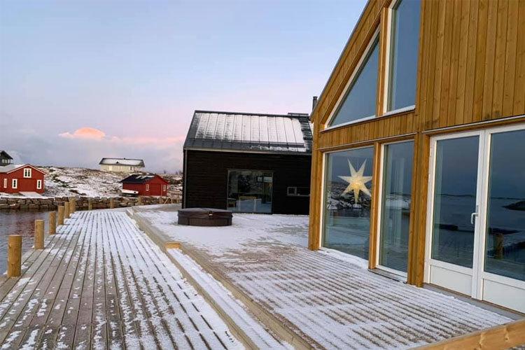 Moderne hytte med store vinduer og tredekk, omgitt av snø og røde bygninger ved vannkanten.