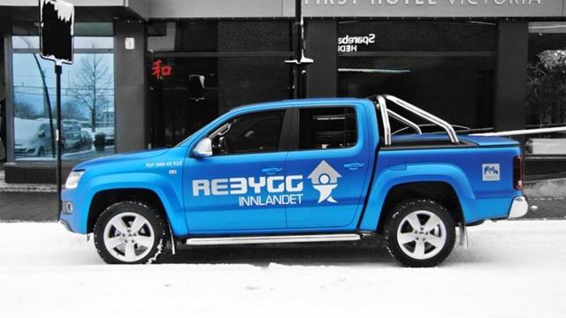 Blå pickup med teksten "REBYGG INNLANDET" parkert på snødekt gate foran hotell.