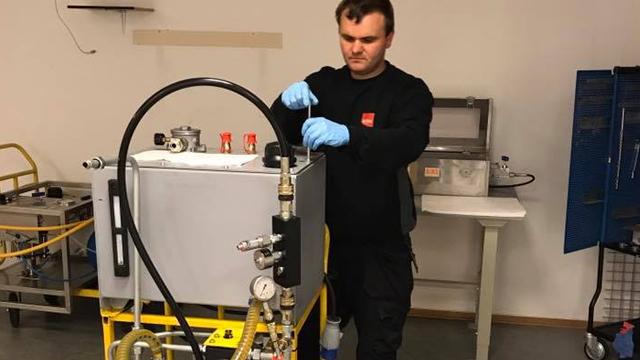 En mann i svart arbeidstøy jobber med en maskin i et laboratorium, iført hansker og blå beskyttelseshansker.