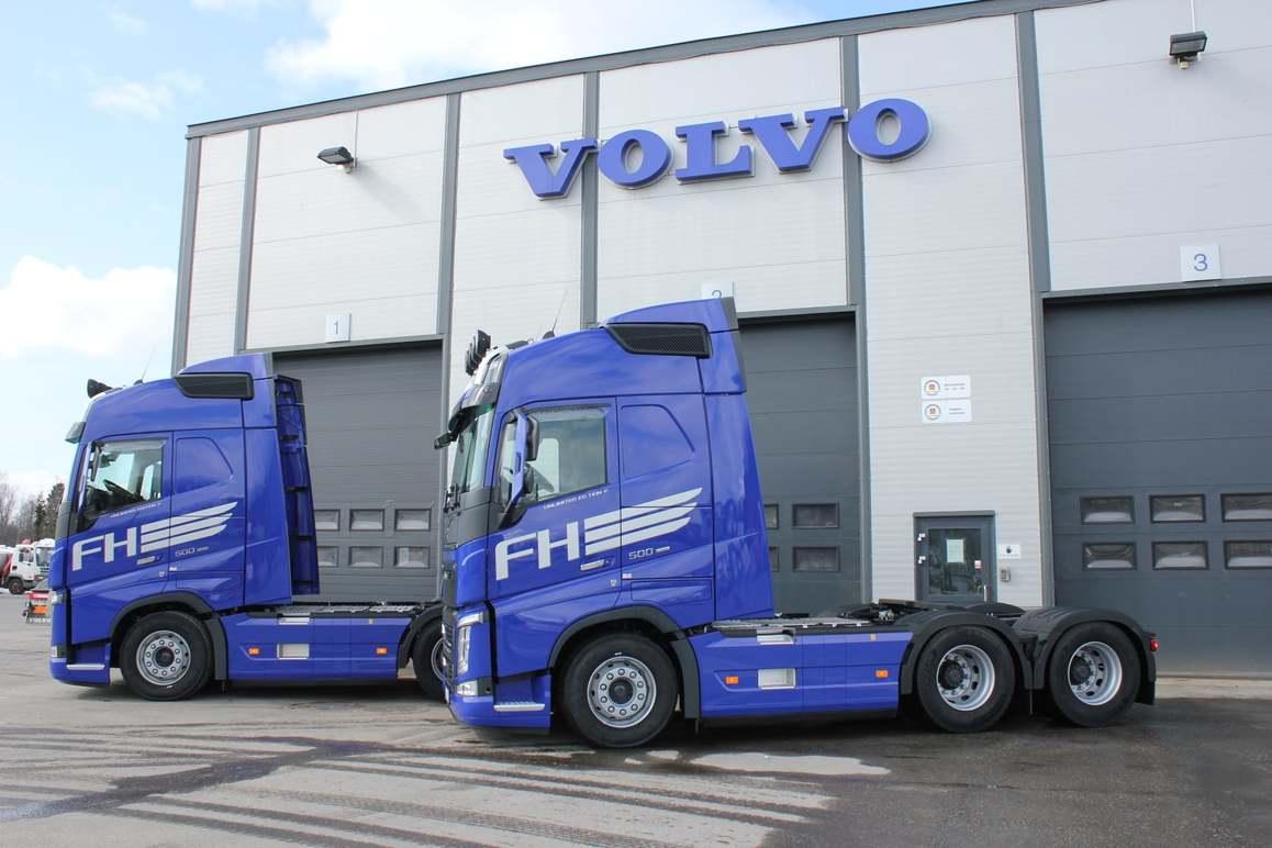 To store med to blå Volvo FH lastebiler parkert foran en bygning med Volvo-logoen.