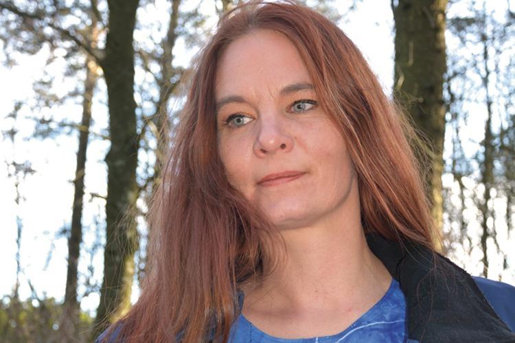 Kvinne med langt, rødt hår står i en skog med trær i bakgrunnen, ser tankefull ut.