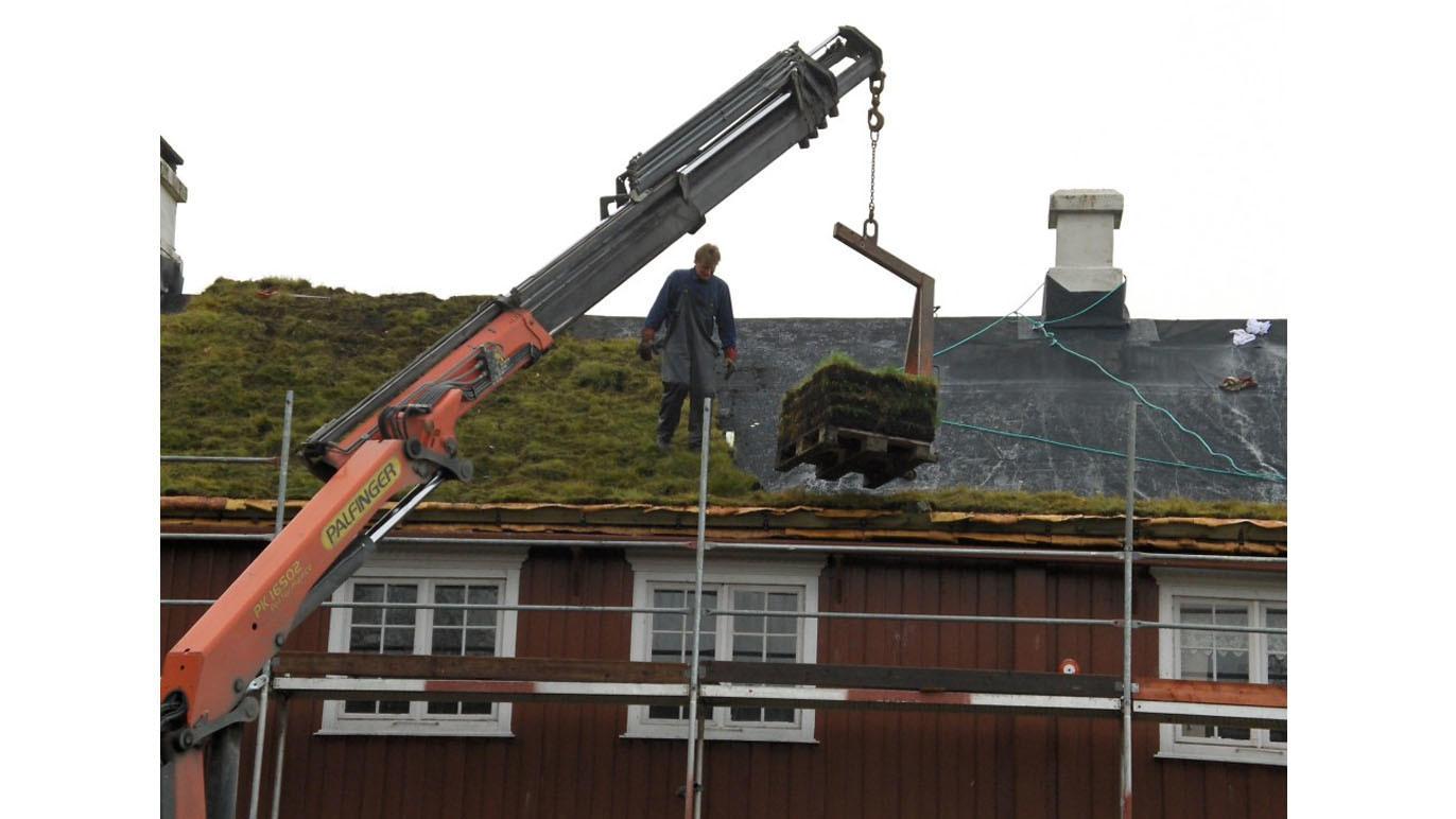 En arbeider fjerner torv fra taket med en kran, mens han står på et stillas ved en bygning.
