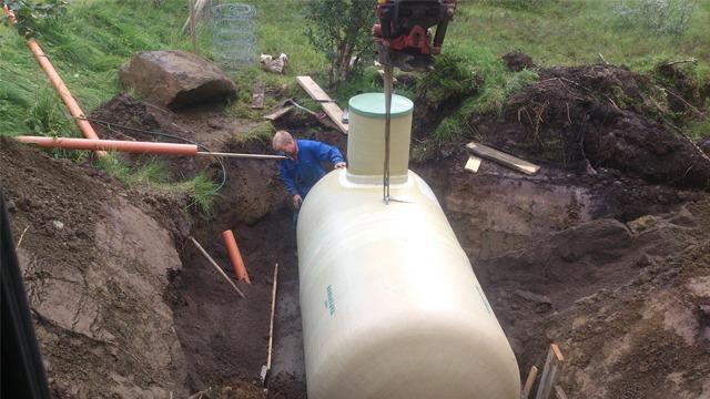 En arbeider installerer en stor tank i en gravd ut grøft, med rør og steiner rundt.