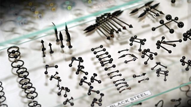 Smykker i svart stål, inkludert piercings og ringer, utstilt på en glassplate.