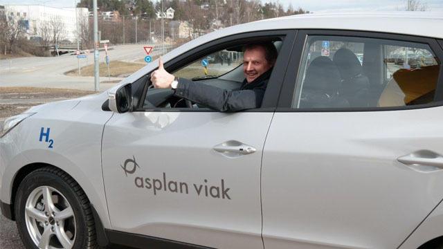 En mann smiler og gir tommel opp fra vinduet på en sølvfarget bil med "asplan viak" logo.