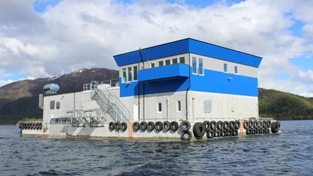 Moderne flytende hus med blå tak og grå vegger, ligger på vannet med fjell i bakgrunnen.