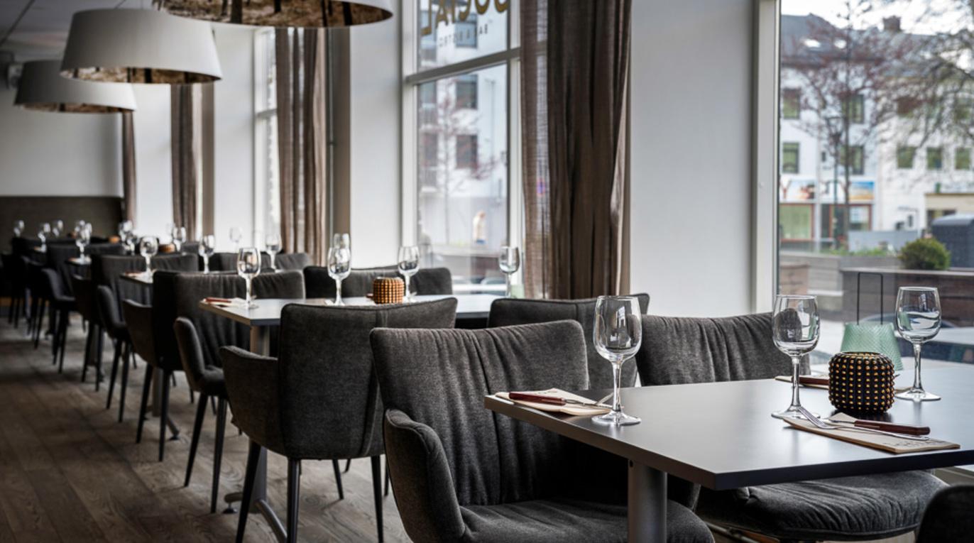 Moderne restaurant med stilige stoler, bord dekket med glass og servietter, og store vinduer som slipper inn naturlig lys.