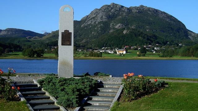 Minnesmerke ved vannet med fjell i bakgrunnen og blomster i forgrunnen.